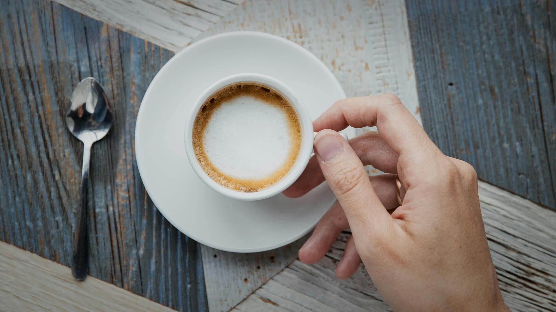 Löffel neben einer gefüllten Kaffee-Tasse mit Cappuccino, welche in einer Hand gehalten wird